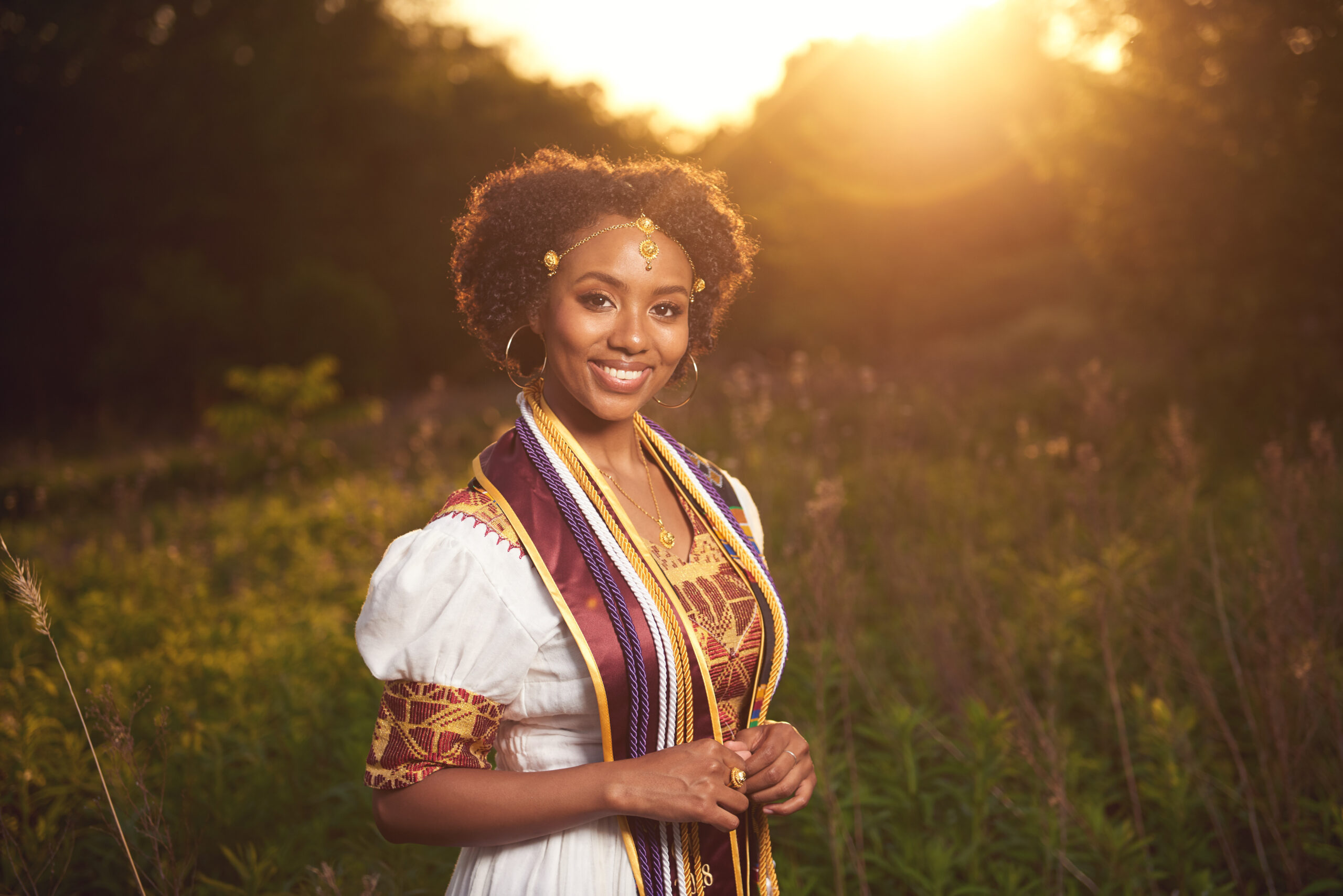 A smiling young woman in traditional attire stands in a beautiful field during sunset, showcasing her culture, proud accomplishments, and joy on the day of her graduation.